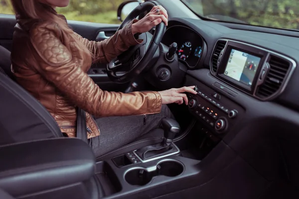 girl car, selects volume level, increases sound in radio, turns temperature control, navigates in car, brown leather jacket, search for route in summer in autumn forest park. Automatic transmission.