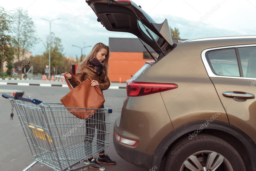 woman at shopping center, opened trunk of a car, puts a shopping bag in trunk of car, brown leather jacket, in summer autumn and spring in city, station wagon, a cart from a store.