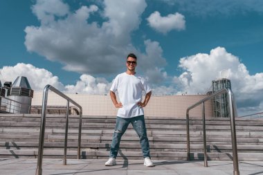Erkek sporcu, yazın şehirde erkek dansçı. Mola dansı için poz veriyorum. Mutlu gülüşler, tişört, kot pantolon ve spor ayakkabı. Arka plan beton merdiven bulutları.