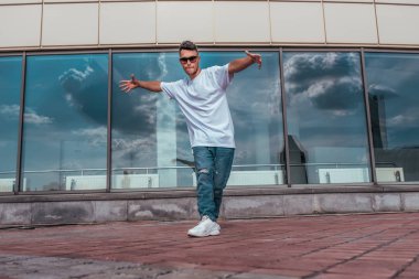 Erkek sporcu, erkek dansçı yaz şehri. Arka plan cam pencere bulutları. Modern break dans tarzı hızlı, fitness sporu hip hop hareketi. Şehir kültürü, sokak dansı. Tişört kotlu spor ayakkabılarıyla..