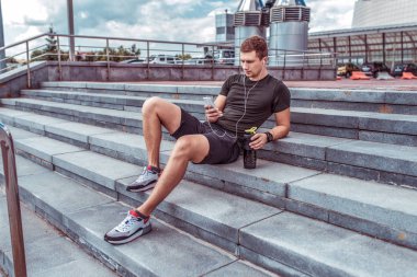 man athlete, summer autumn and spring city, resting active training, holding telephone listening music headphones, watching videos, reading message, active fitness lifestyle, outdoor sports exercise.