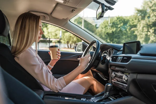 La muchacha a la moda de estilo en el coche, en verano la mujer de la ciudad tiene teléfono móvil, la taza con el té de café, descansa en el estacionamiento, esperando en el atasco de tráfico en la carretera. Snack y almuerzo mientras viaja . — Foto de Stock