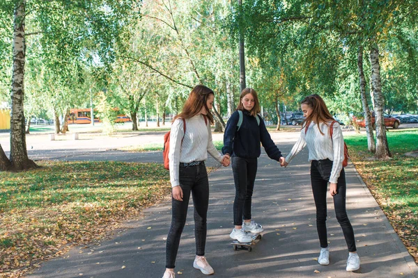 Tonårstjejer i tröjor, 3 skolflickor flickvänner, sommar på hösten och våren i staden, skateboard glad leende ha kul, helg efter skolan. Bakgrund träd gräs, park. — Stockfoto