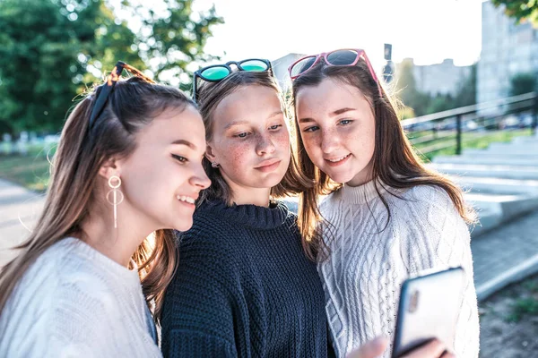 3 девочки-подростки 12-15 лет летом в городе смотреть мобильный телефон, делать селфи, читать смотреть видео приложение онлайн Интернет. Выходные после уроков в колледже. Веселые и смешные . — стоковое фото