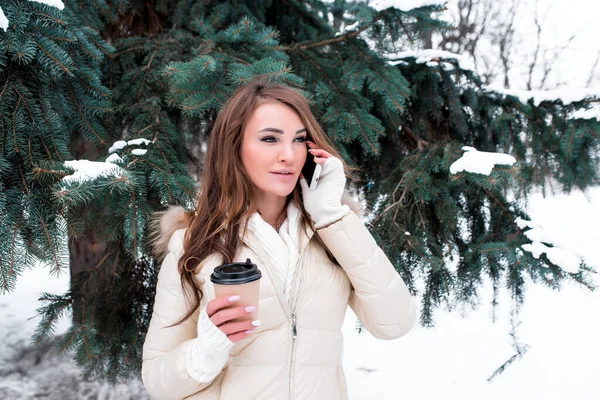 Beautiful girl warm white jacket, winter snowy park, background spruce white snow, cup warm tea coffee calls mobile phone, speaks listens message online Internet application, winter resort weekend. — Stock Photo, Image