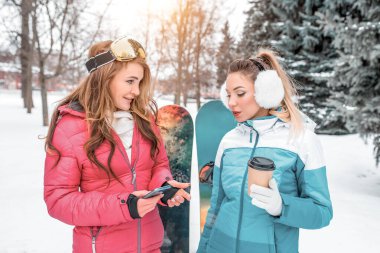 İki kız arkadaş, dinlenme yeri, arka planda kar yağan Noel ağaçları kahve makineleri, dağdan kayak yapmak için kayak tahtası. Spor kıyafetleri spor tulumlarıdır..