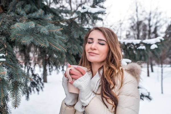 Meisje warme jas, winter achtergrond van sparren sneeuw. Vrije ruimte tekst. Emogenot genot, opwarmt met mok hete koffie thee drinken, dromen glimlacht fantaseert. Lang haar warme wanten. — Stockfoto