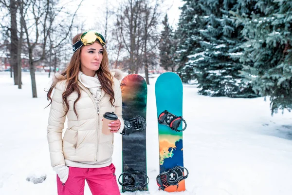 Girl in sportswear, in winter at resort, background snow spruce, snowboard for skiing, cup with coffee tea, glasses for skiing at ski resort. Free space for text. — Stock Photo, Image