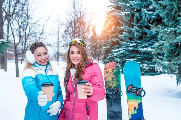 Due ragazze amiche offrono tazze con tè al caffè, si riposano nella località invernale, sullo sfondo di alberi di Natale innevati, tavole da snowboard per sciare dalla montagna. Abbigliamento sportivo è tuta per lo sport . — Foto Stock