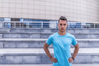 Erkek sporcu, yaz şehrinde, kendinden emin bir şekilde duruyor, koç egzersizi kontrol ediyor. Metin için boş alan. Aktif yaşam tarzı, spor, temiz hava. Spor için motivasyon. Mavi merdiven tişörtü.
