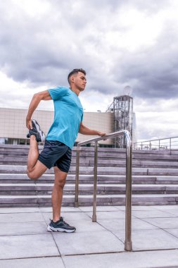 Erkek sporcu, ilkbaharda şehirde koşudan önce bacak ve diz kaslarını ısıtır. Aktif yaşam tarzı, spor, temiz hava. Spor için motivasyon. Tişört şortlar spor ayakkabılar.