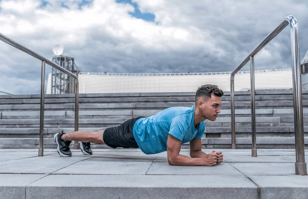 Erkek sporcu, yaz kentinde bahar mevsimi barda duruyor, yalan söylemeyi vurguluyor, karın kasları üzerinde egzersiz yapıyor. Aktif yaşam tarzı, spor, temiz hava. Spor için motivasyon. Tişört şortlar spor ayakkabılar.