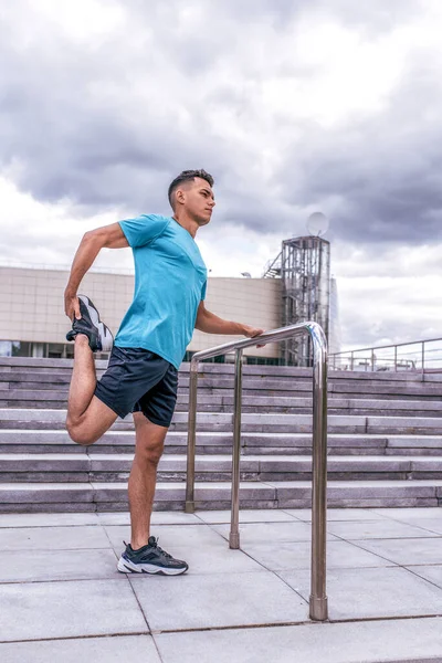 male athlete, summer in spring in city heats up muscles of legs and knee before jogging training. Active lifestyle, workout, fitness in fresh air. Motivation for sports. T-shirt shorts sneakers.