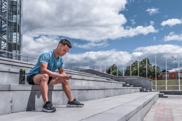 male athlete listens music on phone, reads writes message, online app on Internet, free space text motivation. In summer spring in city. Active lifestyle, workout, fitness in fresh air.