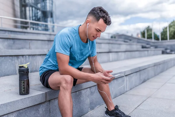 male athlete listens music on phone, reads writes message, an online application on Internet, motivation. In summer and spring in city. Active lifestyle, workout, fitness in fresh air sports.