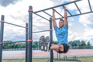 Erkek sporcu, karın kaslarını çalıştırıyor, basın üzerinde egzersiz yapıyor, bacak direğini kaldırıyor. Yazın şehirde. Aktif yaşam tarzı, egzersiz, fitness temiz hava. Motivasyon Mavi tişört, bulut arkaplan.