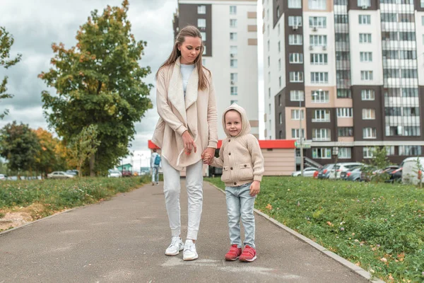 Ibu muda wanita, berjalan di musim gugur dengan anak kecil, anak 3-5 tahun, latar belakang bangunan tinggi, aspal jalan, rumput hijau, pakaian hangat sweater beige . — Stok Foto