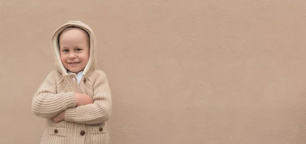 Menino 3-5 anos, na primavera e inverno no verão, está pano de fundo da parede bege, em suéter e capuz. Sorrisos felizes, se alegra e se diverte. Espaço livre para texto. Fundo de Natal . — Fotografia de Stock