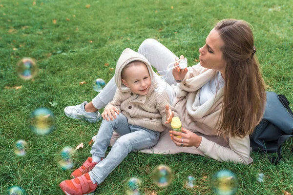 Malý chlapec 3-5 let syn, chytá vzduch a mýdlové bubliny. Máma žena hraje a nafukuje bubliny, happy play baví, na podzim na zelené trávě v teplém oblečení. — Stock fotografie