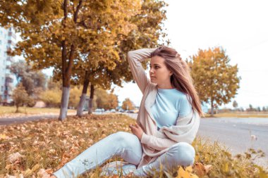 Sonbaharda kadın yapraklarla çimenlerde oturur, arka planda ağaçlar yol, dinlenme bakışları, uzun saçlar, kalın elbiseler, bej pantolon..