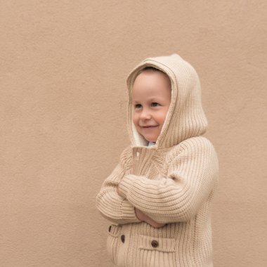 3-5 yaşlarında küçük bir çocuk, ilkbahar ve kışın, bej bir duvarın arkasında duruyor, kazak ve kapüşonlu. Mutlu gülücükler, neşe ve eğlence..