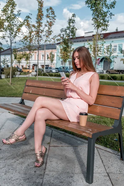 Estudiante con un vestido rosa, en verano en la ciudad se sienta en un banco, lee y escribe un mensaje en un teléfono móvil, una aplicación en línea en Internet. Redes sociales, una taza de té de café . —  Fotos de Stock