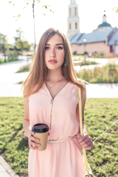 Девочка летом в розовом платье, рука в чашке с напитком, длинные волосы, яркий солнечный день. Каждый день макияж. Выходные будут спокойными и комфортными . — стоковое фото