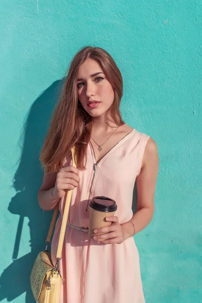 Siswa perempuan dengan gaun merah muda, di musim panas di kota, dinding latar belakang, cangkir dengan teh kopi, tas kuning, rambut panjang, riasan kasual . — Stok Foto