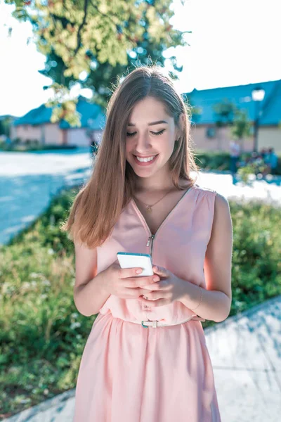 Chica estudiante en vestido rosa, en el parque de la ciudad de verano feliz sonriente se regocija y ríe, árboles de fondo arbustos, lee y escribe mensaje en el teléfono móvil, aplicación en línea en Internet . —  Fotos de Stock