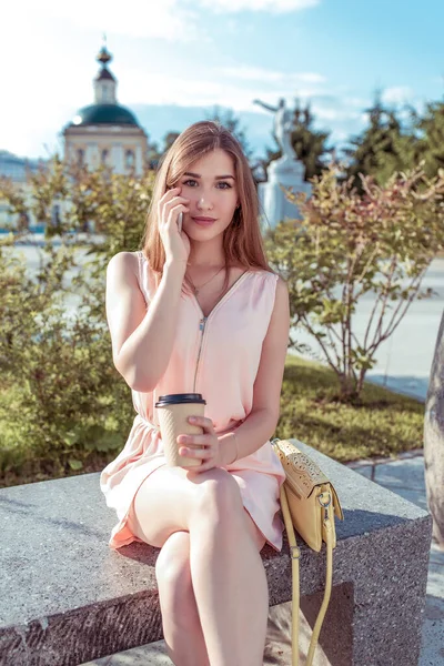 Chica estudiante en vestido rosa, parque de la ciudad de verano se sienta banco, árboles de fondo arbustos, hace llamada telefónica escucha el mensaje en el teléfono móvil, aplicación en línea café taza de Internet con té, bolso amarillo . — Foto de Stock