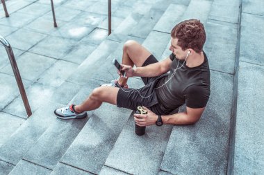Atletik adam yazın merdivenlerde dinleniyor. Spor yaptıktan sonra dışarıda spor yapıp dinlenin. Telefonun elinde, internette müzik uygulaması dinliyor. Su ve proteinli titretici..
