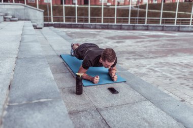 Yazın şehirde sporcu, barda duruyor, karın kaslarını ve basını eğitiyor, yoga minderi, telefon uygulaması online akıllı telefon zamanlayıcısı, su proteiniyle karıştırıcı.