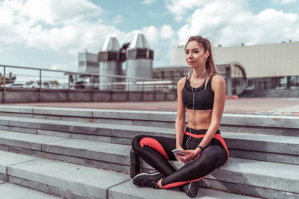 Atlet kvinne hviler om sommeren på trappene by. Hvile etter trening og utendørstrening. Han lytter til musikkprogrammer på nettet. Fritt rom for tekst . – stockfoto