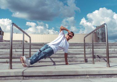 Tek kollu dansçılar break dans, hip-hop. Yazın, şehrin arka planında bulutlar dolaşıyor. Aktif gençlik yaşam tarzı, genç erkek dansçı, fitness hareketi break dans kot ayakkabıları, güneş gözlüğü..