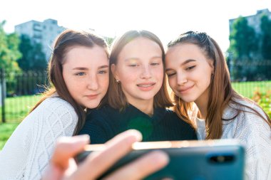 Üç kız öğrenci kız mutlu bir şekilde gülümsüyor ve akıllı telefondan fotoğraf çekiyor. Telefondaki selfie 'ler, sosyal ağlar, online uygulama, internete video çağrısı. Yazın doğada.