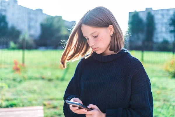 Gadis remaja sekolah, latar belakang kota musim panas adalah rumput hijau, sweater hangat, memegang ponsel, membaca dan menulis pesan, aplikasi online di jejaring sosial Internet . — Stok Foto