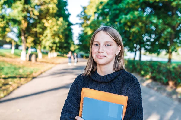 Meisje tiener schoolmeisje, zomer stad achtergrond bomen en weg, warme trui, in handen van notebooks, glimlachen blij, na lessen op school en instituut, vrije ruimte voor tekst. — Stockfoto