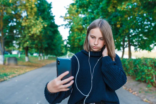 10代の女の子の女子高生ヘッドフォンで写真を撮る10代の女の子自身の電話、真剣な女の子は会話、録音、ビデオ通話、秋の夏の都市、黒いセーターを聞きます。背景木道. — ストック写真