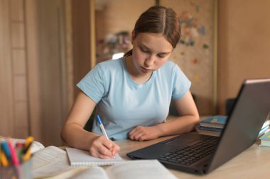 Teen girl studying at home with a laptop, writing in a notebook, education at home, preparing for the exam at the institute and school. Qualification courses, video conference on the Internet.