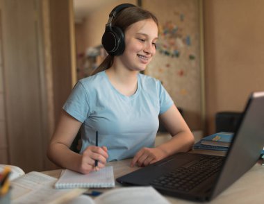 Happy teenager girl smiling on video call with teacher and parent, internet conference, home education and learning to school and college. Headphones notepad and notebook writing.