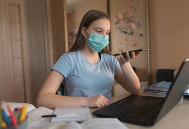 Teenager girl, home lessons laptop, medical mask, protection bacteria and viruses, talking on smartphone, recording audio call, video tutoring lessons, home education video conference on Internet.