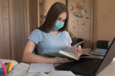 Teenager girl, home lessons at a laptop, medical mask, protection against bacteria viruses, reads a paper book textbook, video tutoring lessons, home education, preparation for college and school.