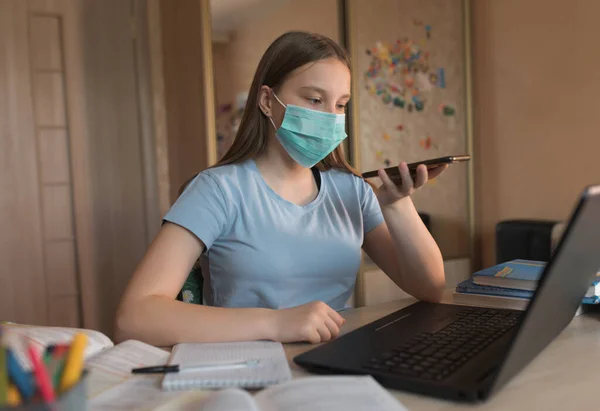 Teenager girl, home lessons laptop, medical mask, protection bacteria and viruses, talking on smartphone, recording audio call, video tutoring lessons, home education video conference on Internet. — Stock fotografie