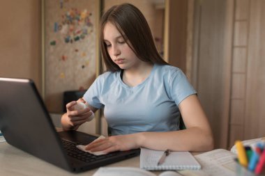 Teenager girl, home lessons at a laptop, antiseptic spray, disinfection of a laptop keyboard from bacteria and viruses, video tutoring lessons, home education, preparation for college and school.