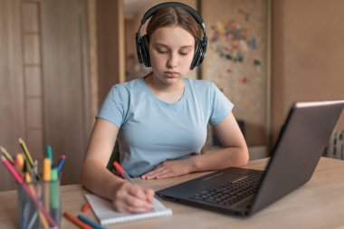 Teen girl doing homework with headphones, laptop e-learning and online lessons. Social network video call, listens to the teachers audio lecture on the Internet.