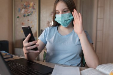 Teenager girl, in a medical mask, protection bacteria of viruses and diseases, makes a video call on a smartphone, e-education and Internet lessons, distance learning. Video training on the Internet.
