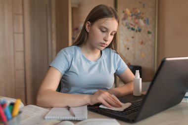 Teen girl rubbing a laptop keyboard with a napkin, protecting bacteria from germs and dust. Personal hygiene, antiseptic spray. E-education and online lessons, distance learning.
