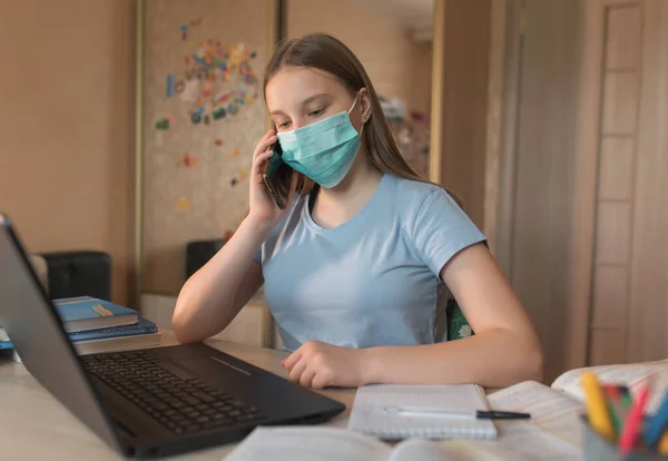 Teenager girl, in a medical mask, disinfection from bacteria and germs and viruses, calls on a smartphone, laptop monitor, e-education and Internet lessons, distance learning. — Stock fotografie