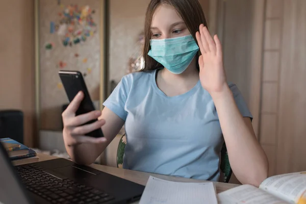 Teenager girl, in a medical mask, protection bacteria of viruses and diseases, makes a video call on a smartphone, e-education and Internet lessons, distance learning. Video training on the Internet. — Stock fotografie