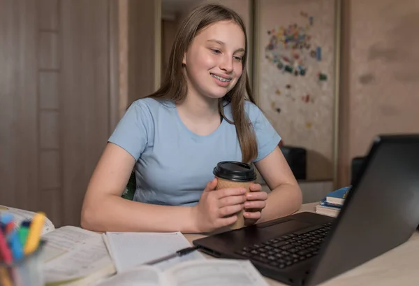 Teen girl, happy smiling, braces on her teeth, watching funny videos on social networks Internet, a break from school. E-education online lessons, distance learning. Notebook cup coffee tea, on home.